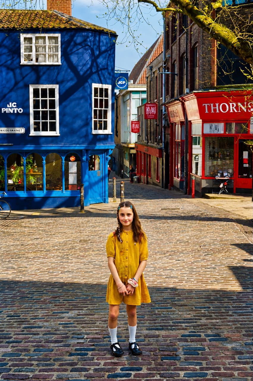 Girl in a yellow dress on a cobbled street between blue and red shopfronts
