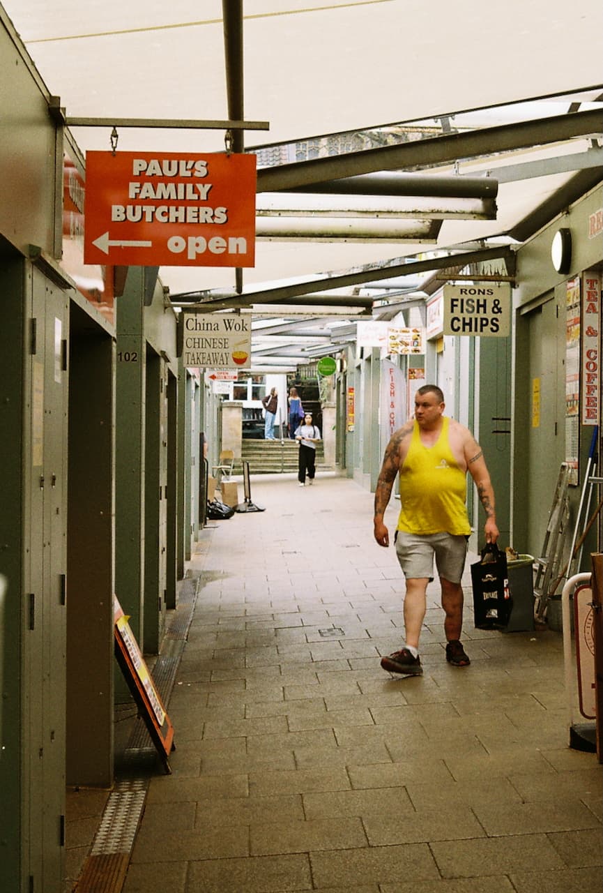 Man in a yellow vest walking through a covered market with food stall signs